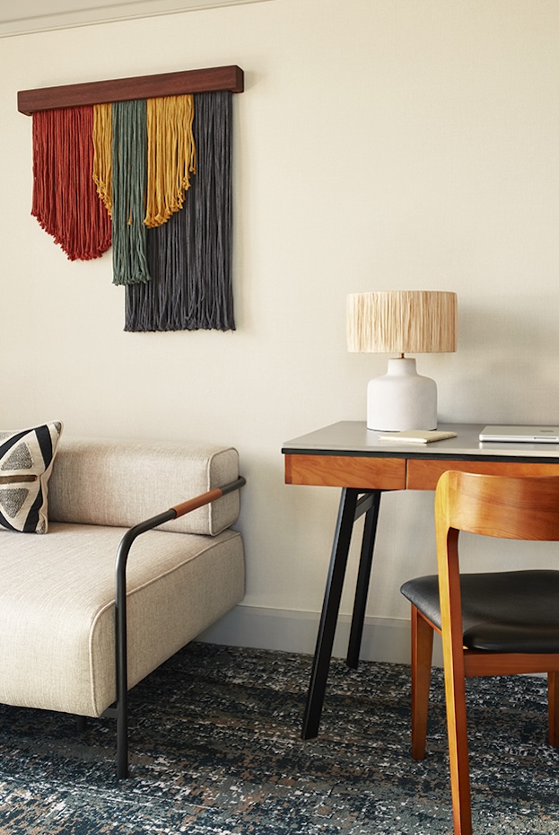 A workspace corner featuring a mid-century modern wooden desk and chair. Above the desk, a colorful fiber art wall hanging in terracotta, forest green, and mustard yellow adds texture to the cream wall. A small white lamp with a textured shade sits on the desk.