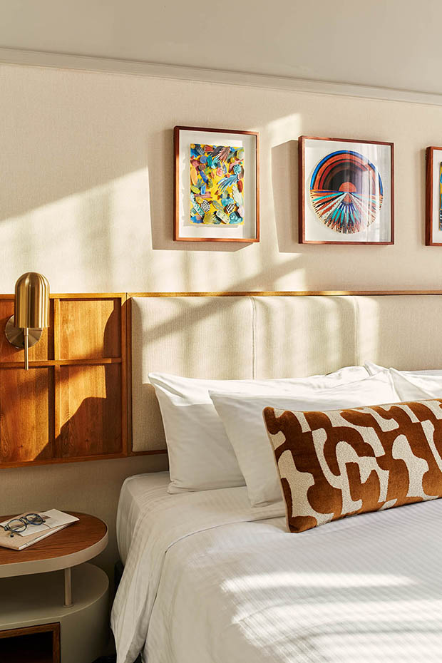 Close-up of a plush king bed with white linens and a brown patterned lumbar pillow. The headboard features integrated wood paneling and a brass sconce, with colorful framed abstract art hanging on the wall above.