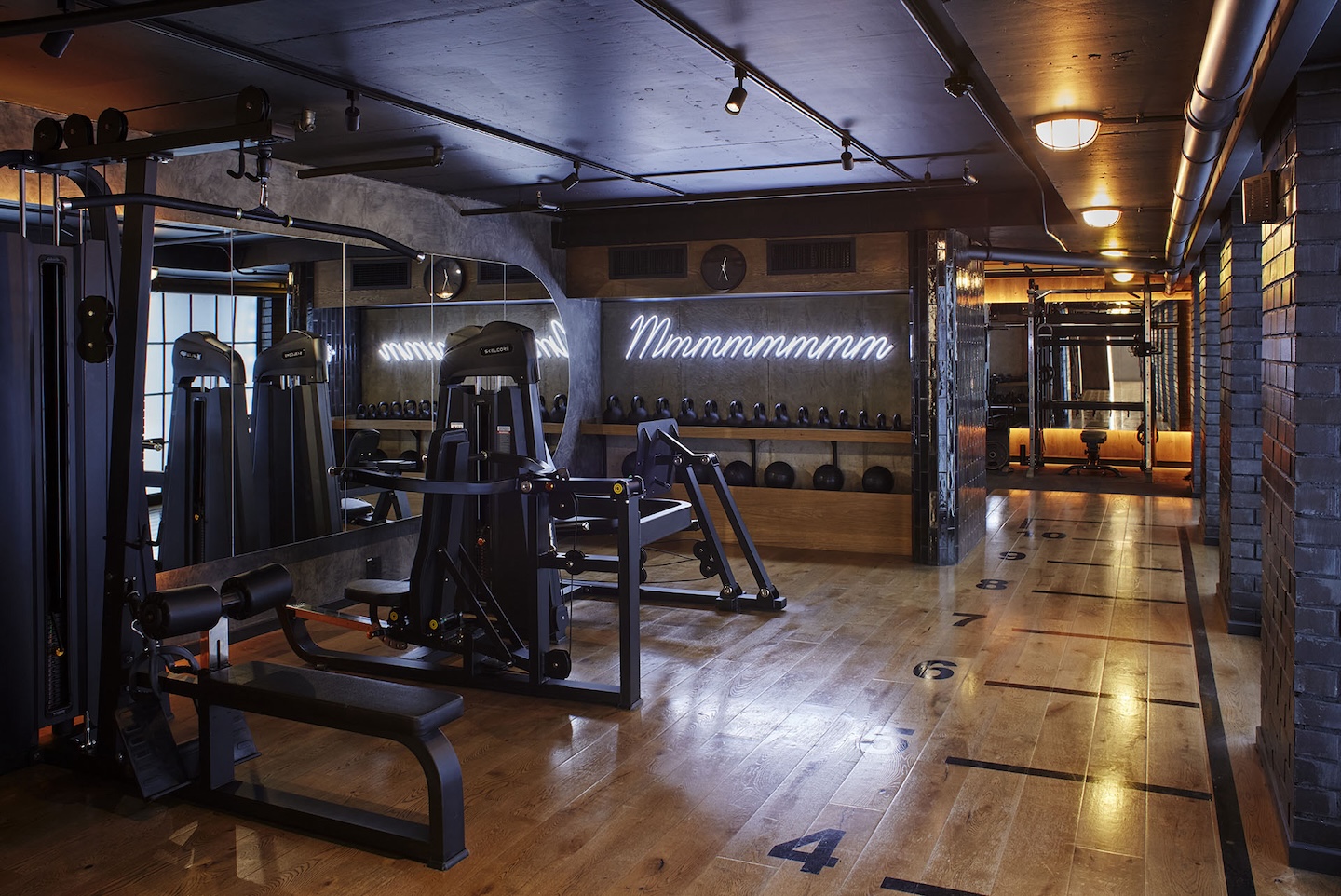 A sleek weight training room featuring various resistance machines and a row of kettlebells and medicine balls on a wooden shelf. A white neon sign that reads "Mmmmmmmm" glows on the concrete wall. The wood floor includes numbered markers for guided workouts.