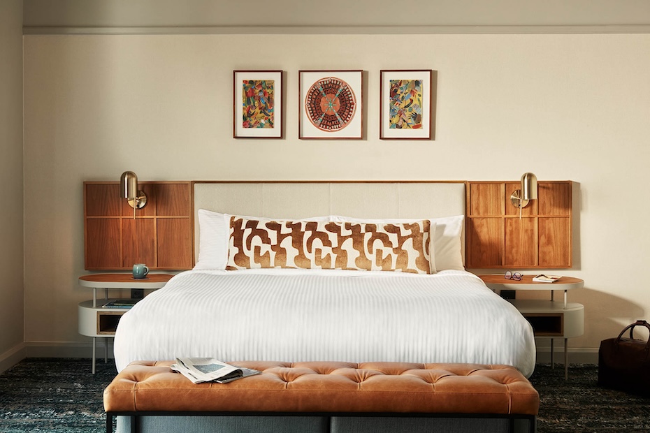 A king-sized bed with crisp white linens and a long, brown-and-white patterned lumbar pillow. The headboard features integrated wood paneling and brass reading lamps. A tan tufted leather bench sits at the foot of the bed, and three framed pieces of abstract art hang on the wall above.