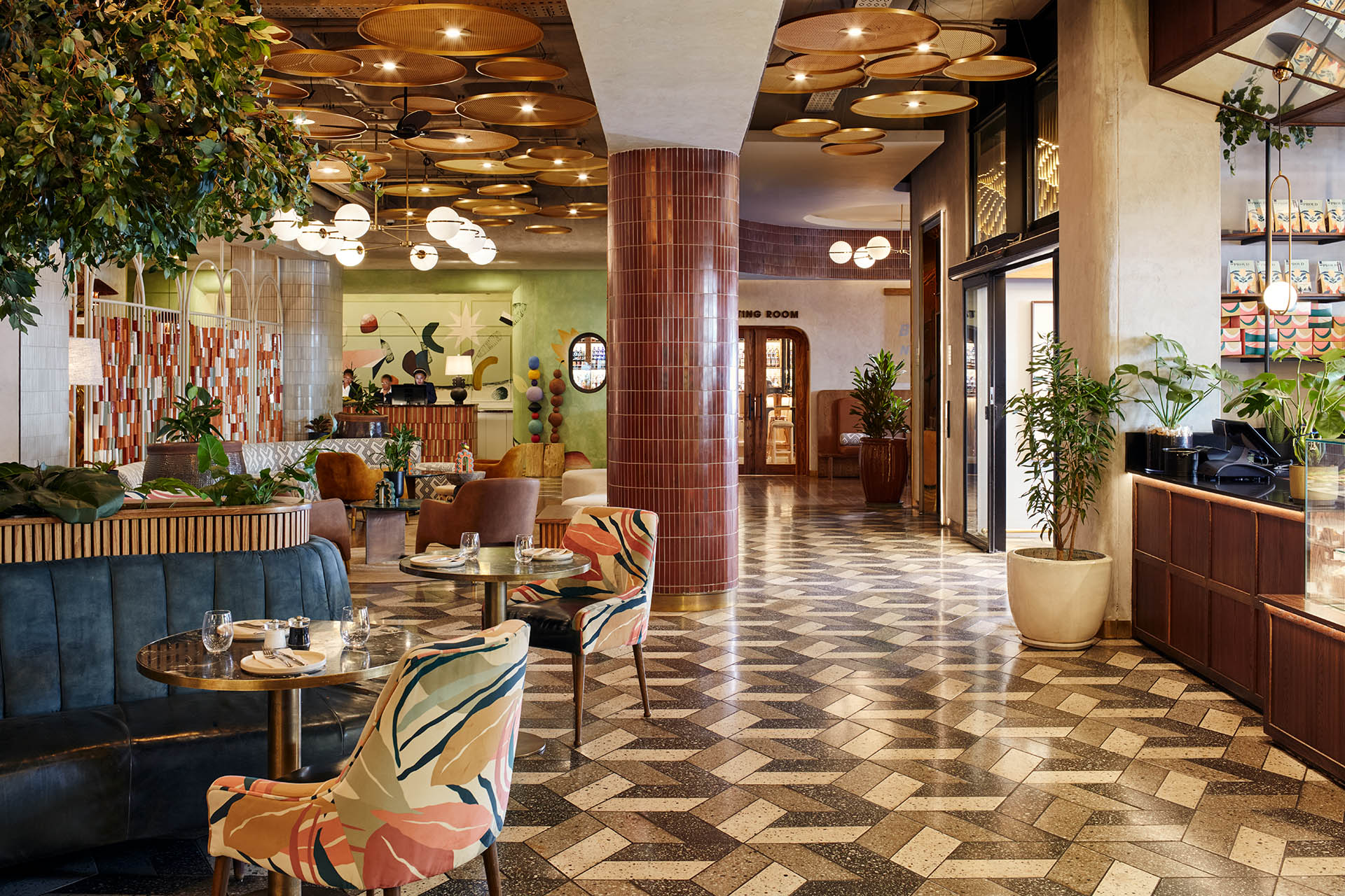 A spacious hotel lobby with a geometric patterned floor, featuring a mix of colorful floral-patterned chairs and velvet banquette seating. The ceiling is decorated with numerous circular woven panels and modern globe lighting.