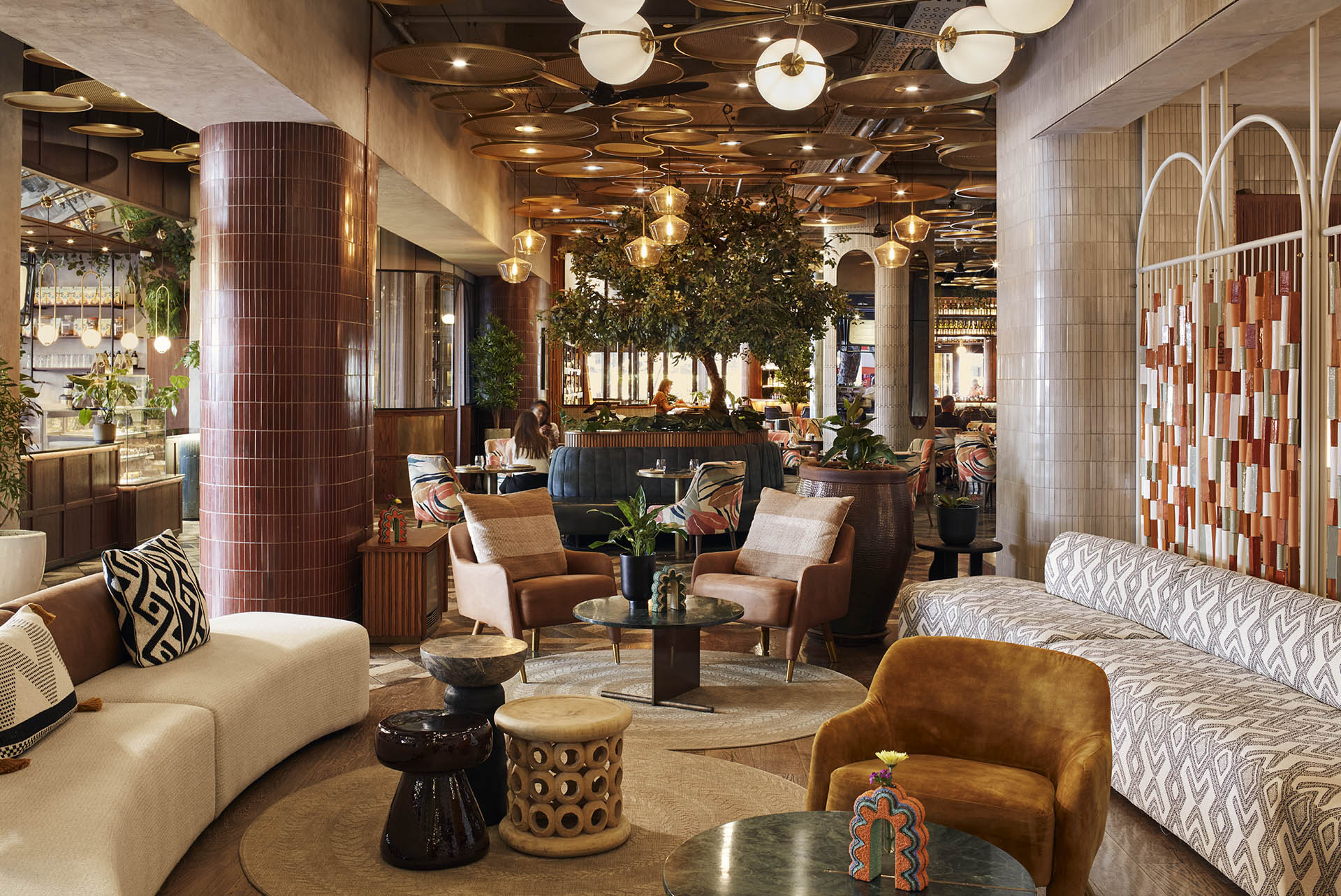 A vibrant hotel lounge featuring a variety of mid-century modern furniture, including leather armchairs and patterned sofas. A large indoor tree serves as a centerpiece, surrounded by warm globe lights and textured pillars.