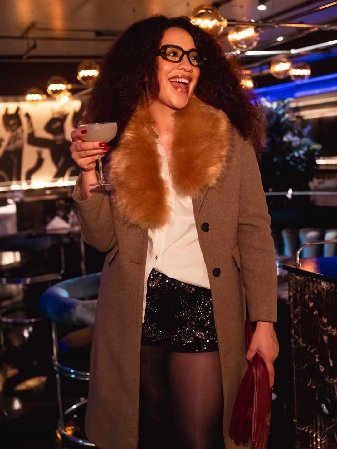 A woman with long curly hair and glasses smiling while holding a cocktail in a coupe glass. She is wearing a brown coat with a large fur collar and sequined shorts in a dimly lit, upscale lounge.