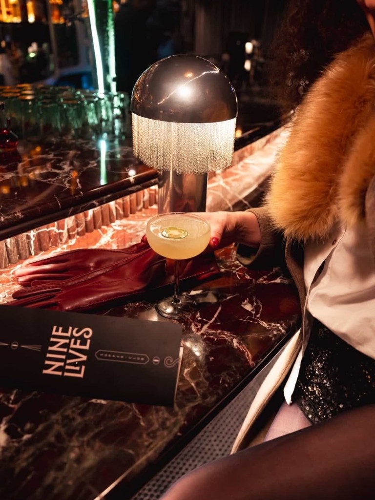 A person in a fur-collared coat reaches for a light-colored cocktail in a coupe glass garnished with a lime slice. A "Nine Lives" menu and a pair of red leather gloves rest on the marble bar nearby.