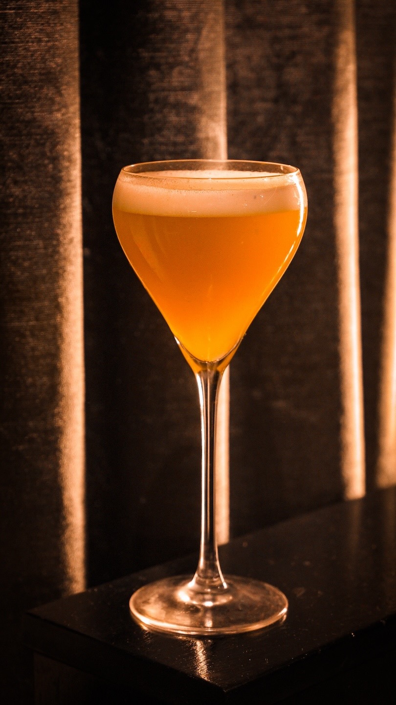 A vibrant orange cocktail served in a tall, elegant Nick and Nora glass with a thin layer of white foam on top. The glass is set against a backdrop of vertical, dark textured curtains under warm, dramatic lighting.
