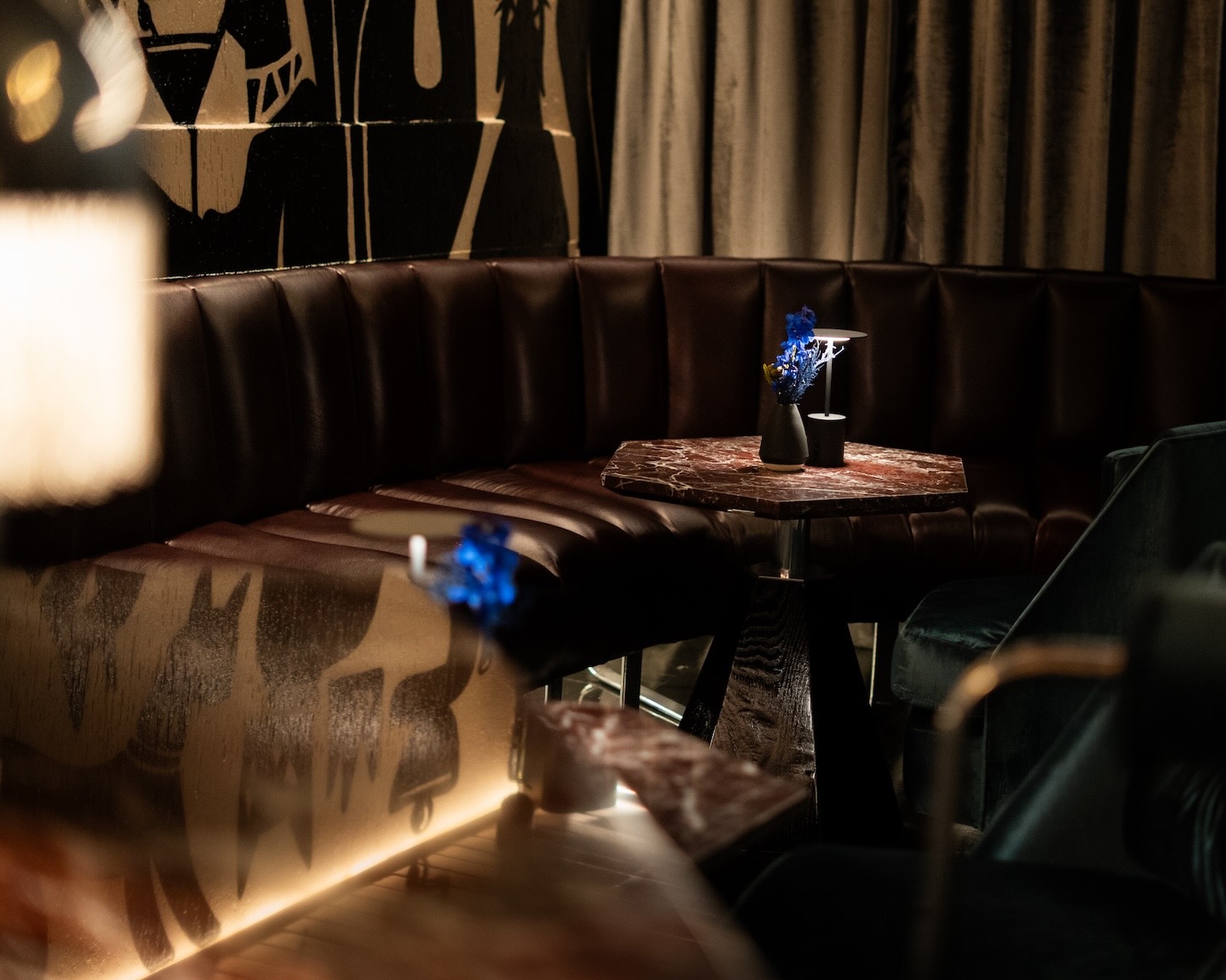 A moody, low-angle shot of a curved brown leather booth in a lounge. A small, hexagonal marble table holds a black vase with blue flowers and a modern lamp, set against a wall with black and white feline-themed murals.