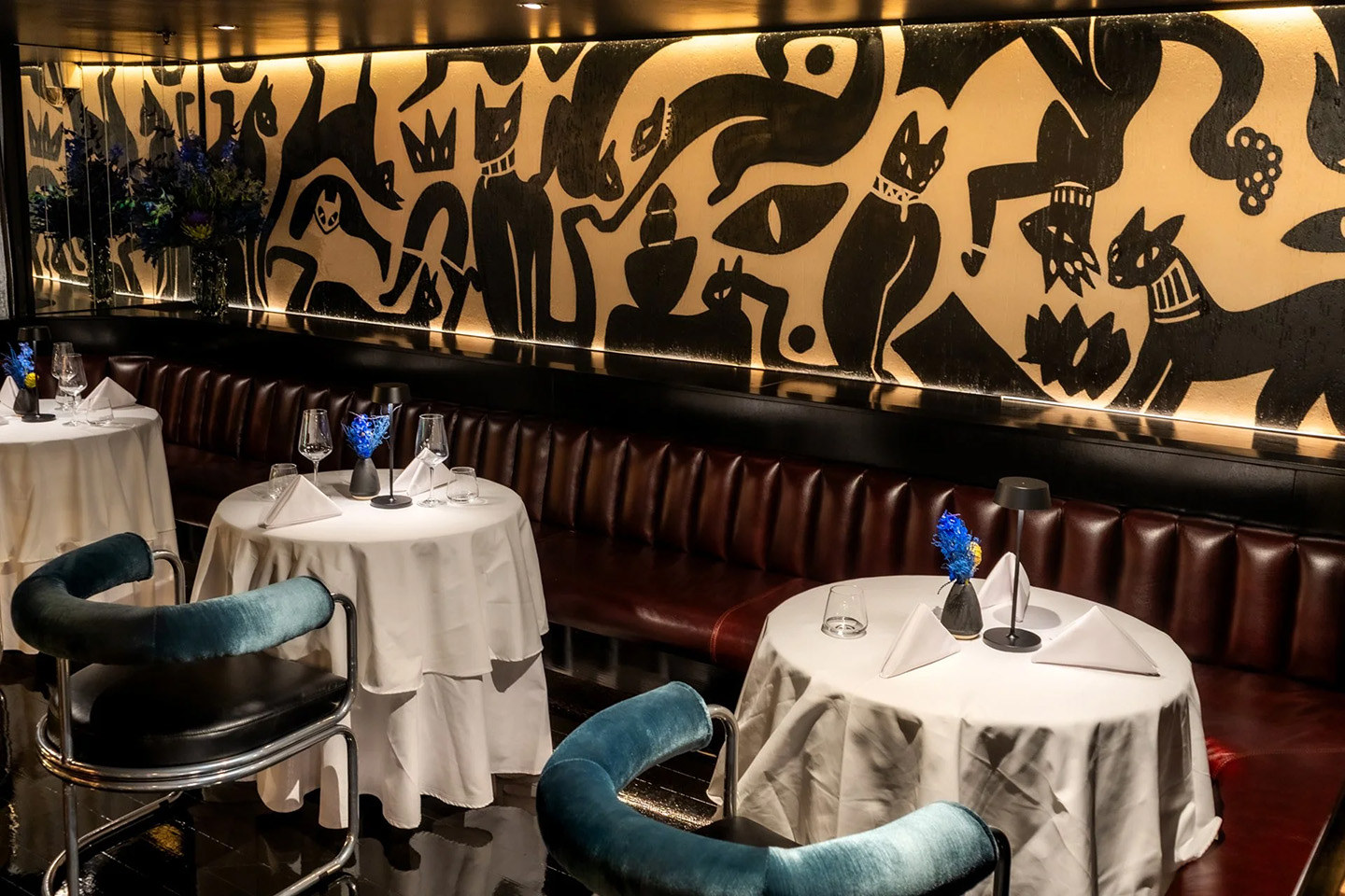 A wide view of a stylish lounge interior featuring dark polished floors, teal velvet chairs, and a long curved leather banquette. A large mural of stylized black cats stretches across the back wall, and a marble-topped bar with leather stools is visible on the left.