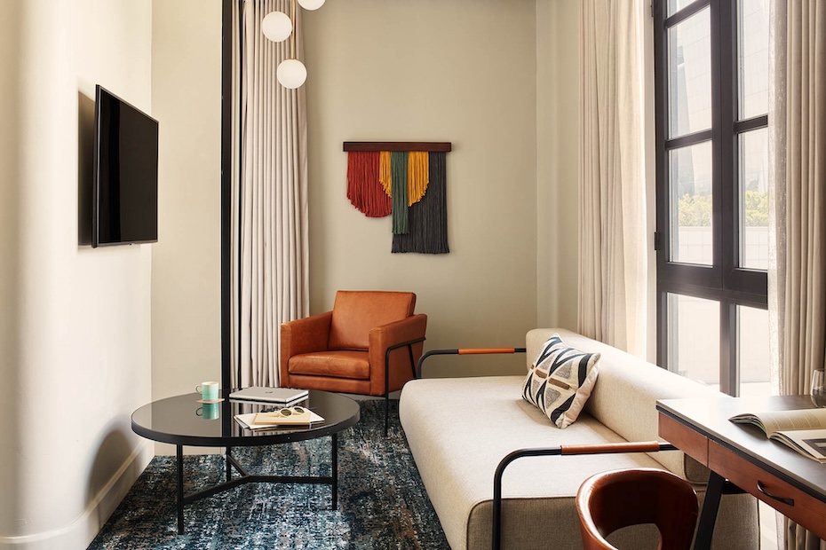 The living area of a hotel suite featuring a beige sofa with a geometric pillow, a black round coffee table, and a burnt orange leather armchair. Large floor-to-ceiling windows with sheer curtains let in natural light, and a colorful fiber art piece hangs on the wall.