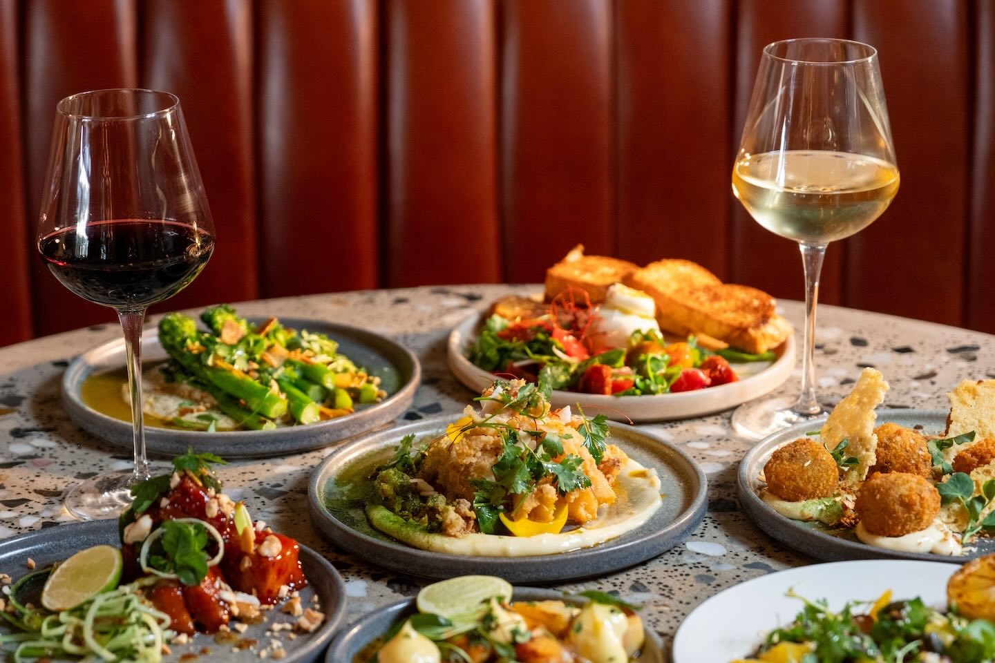 A wide shot of a dinner table featuring several shared plates, including glazed pork belly, charred broccoli, and arancini. A glass of red wine and a glass of white wine are positioned on opposite sides of the frame.