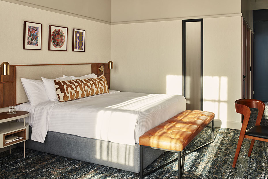 Wide shot of a king-sized bed with a neutral upholstered headboard and integrated wooden nightstands with brass reading lamps. Three pieces of colorful framed art are mounted above the bed. Sunlight creates sharp geometric shadows across the bed linens and the tan leather bench.